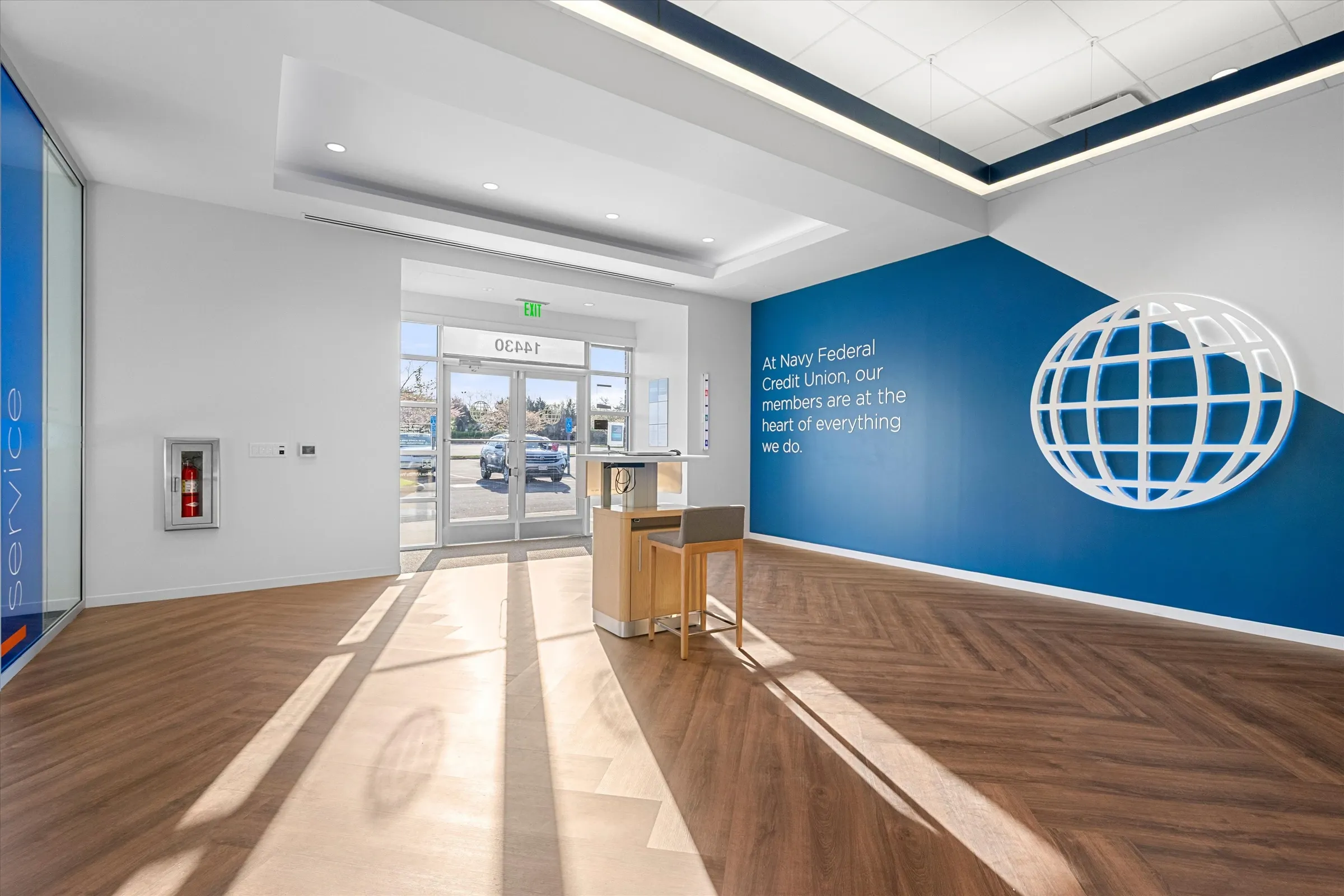 Branch entry foyer with herringbone wood flooring, branded blue mission wall with 3D globe logo, glass doors, and greeter podium