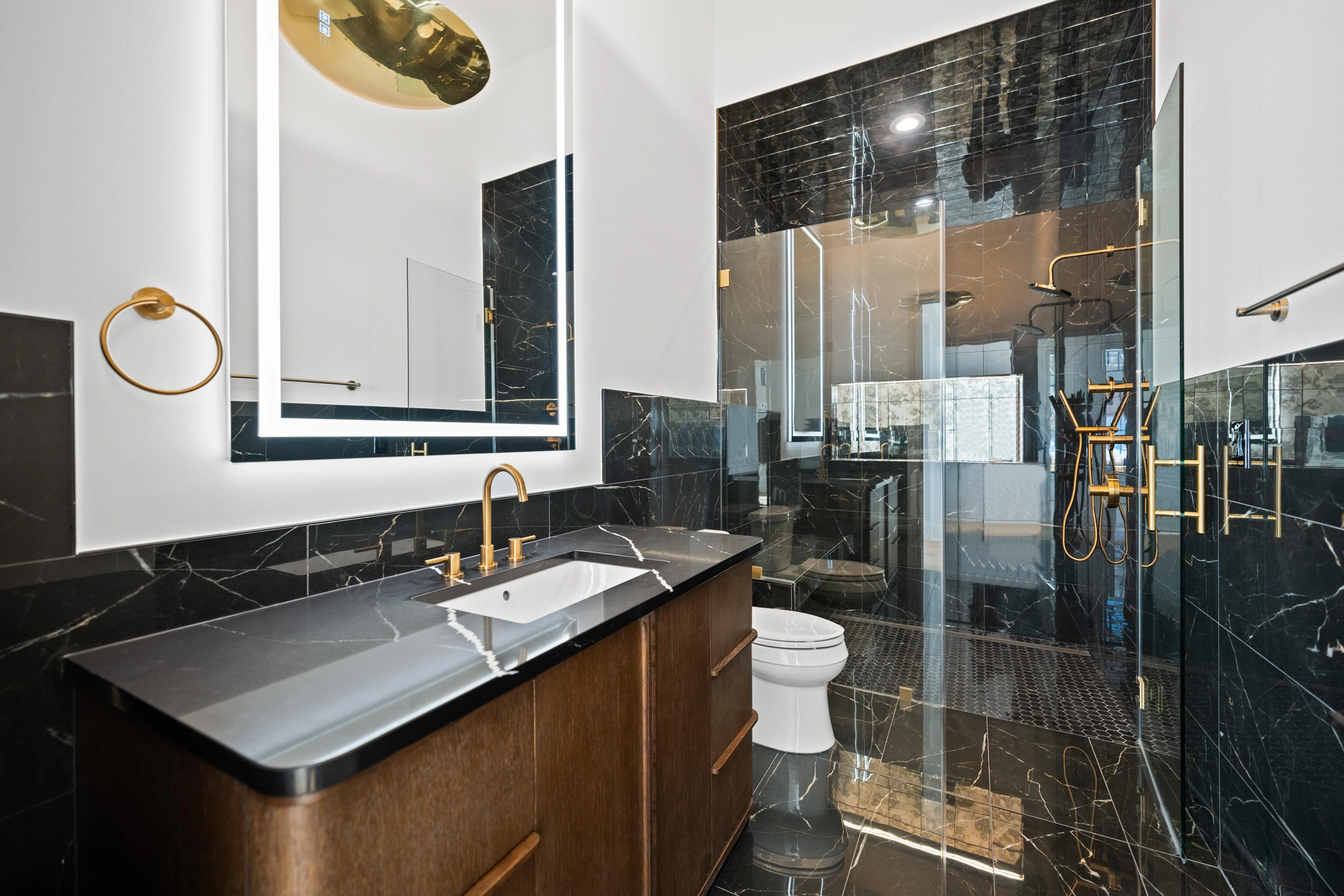 Dramatic en-suite bathroom with black marble walls, gold fixtures, LED backlit mirror, and glass shower enclosure
